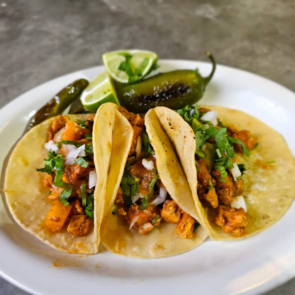 Three Los Agaves Taqueria tacos with meat, cilantro, and onions, served with limes and jalapeños.